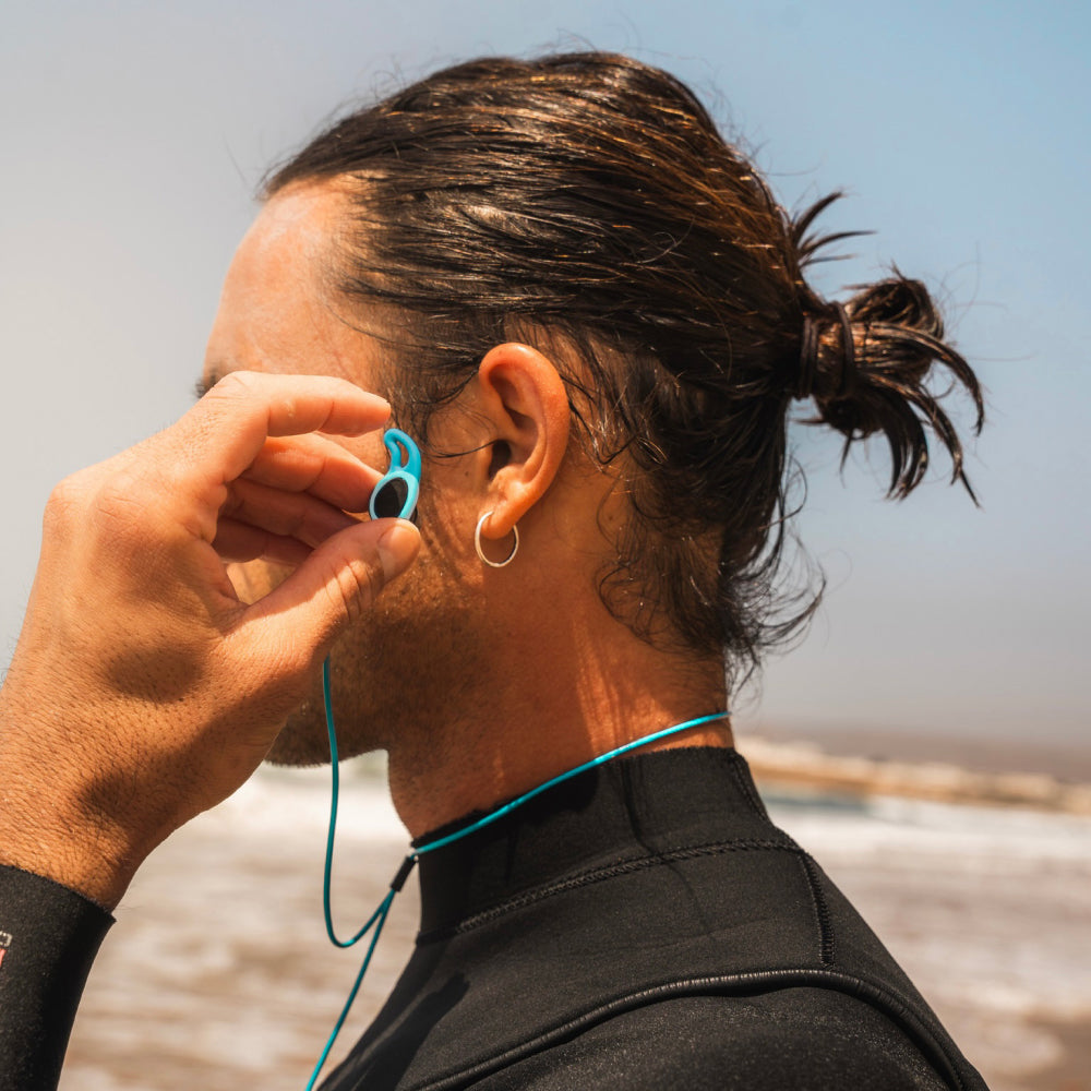 Man inserting blue water-sports earplugs while standing by the ocean.